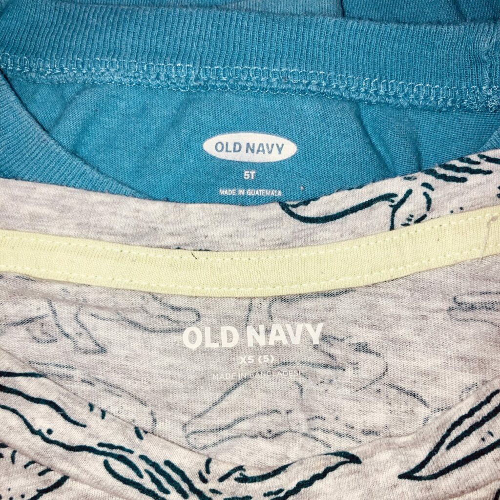 Clothing tags showing 5T vs. XS sizes on two Old Navy shirts. The photo is zoomed in on the neck opening of the shirts. The back shirt is blue and the front shirt has an alligator print.