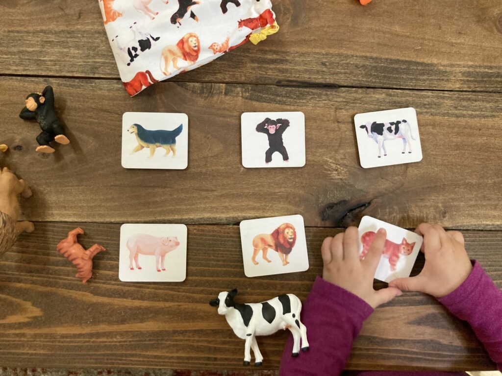 Lovevery wooden animal tiles with animal figurines (cow, cat, monkey) and baby hands picking up a tile