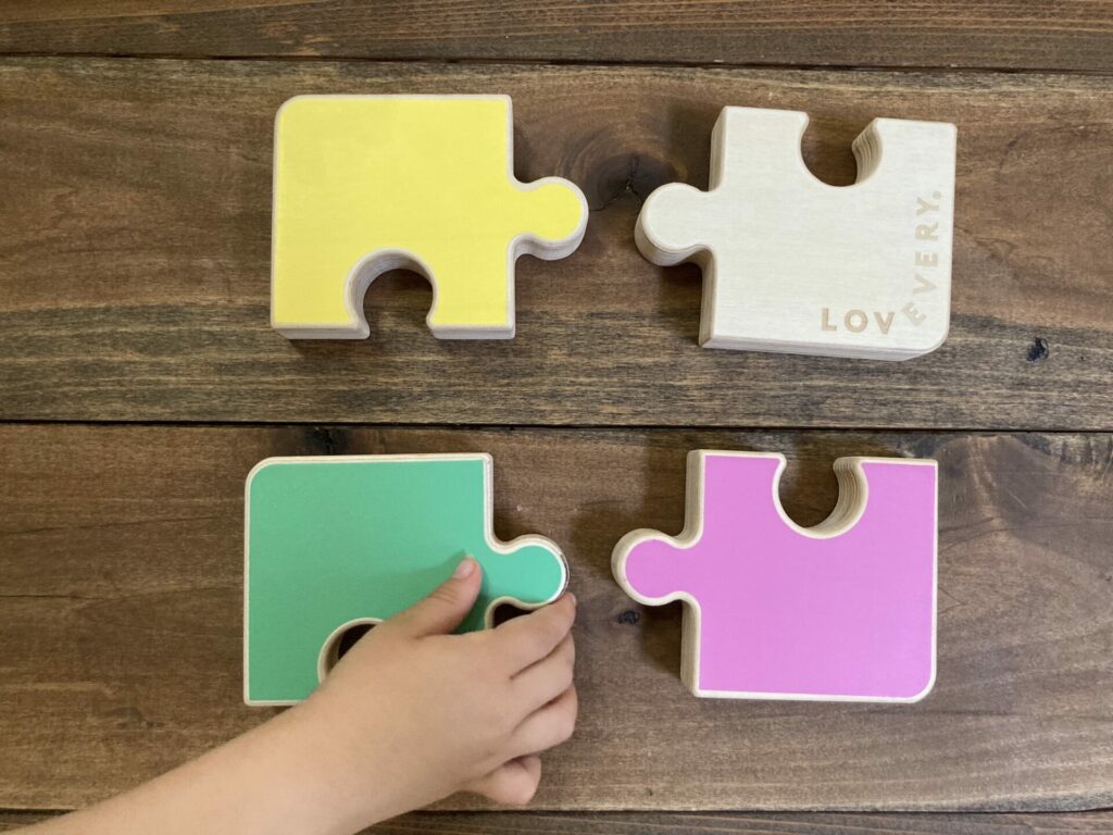 chunky lovevery puzzle, showing 4 colorful puzzle pieces and a baby hand reaching for the green piece