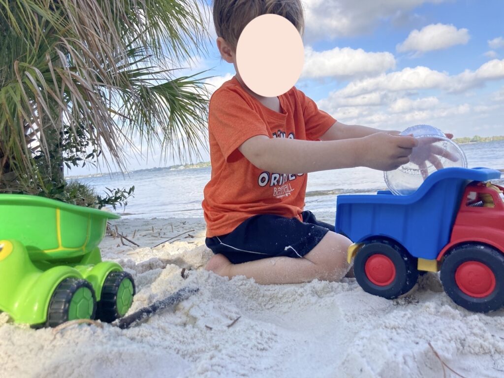 kid with this face obscured playing with a blue and a green dump truck at the beach.