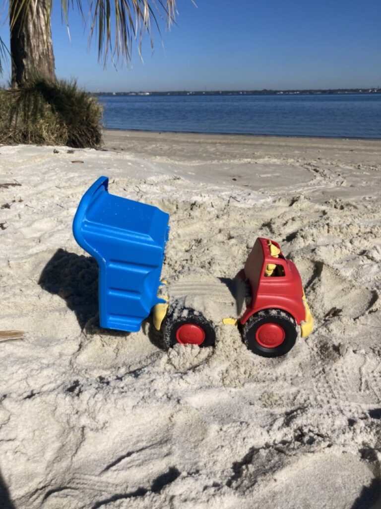 green toy dump truck with the dump open and the truck partially buried in the sand. the truck is red and the dump is blue