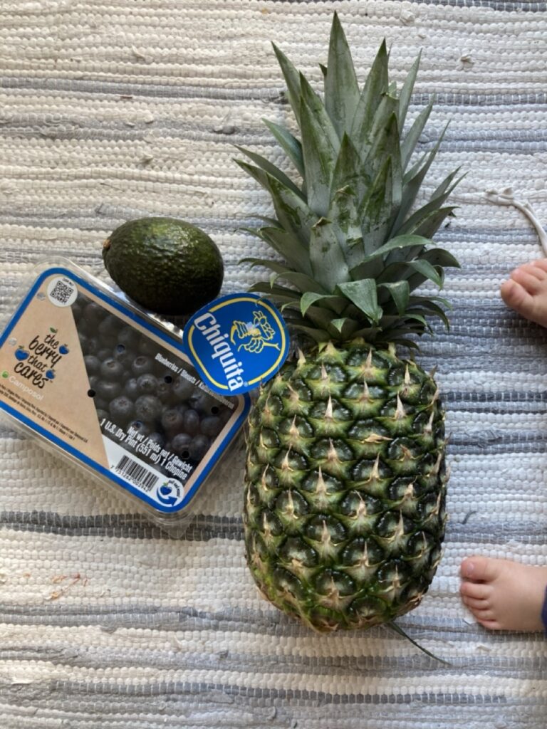 A pineapple, blueberry container, avocado on the floor with baby feet on the edge of the frame
