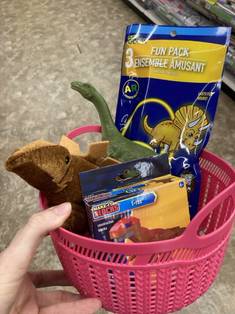 This picture is of a small plastic basket filled with dinosaur toys being held by a hand in an aisle at the dollar store