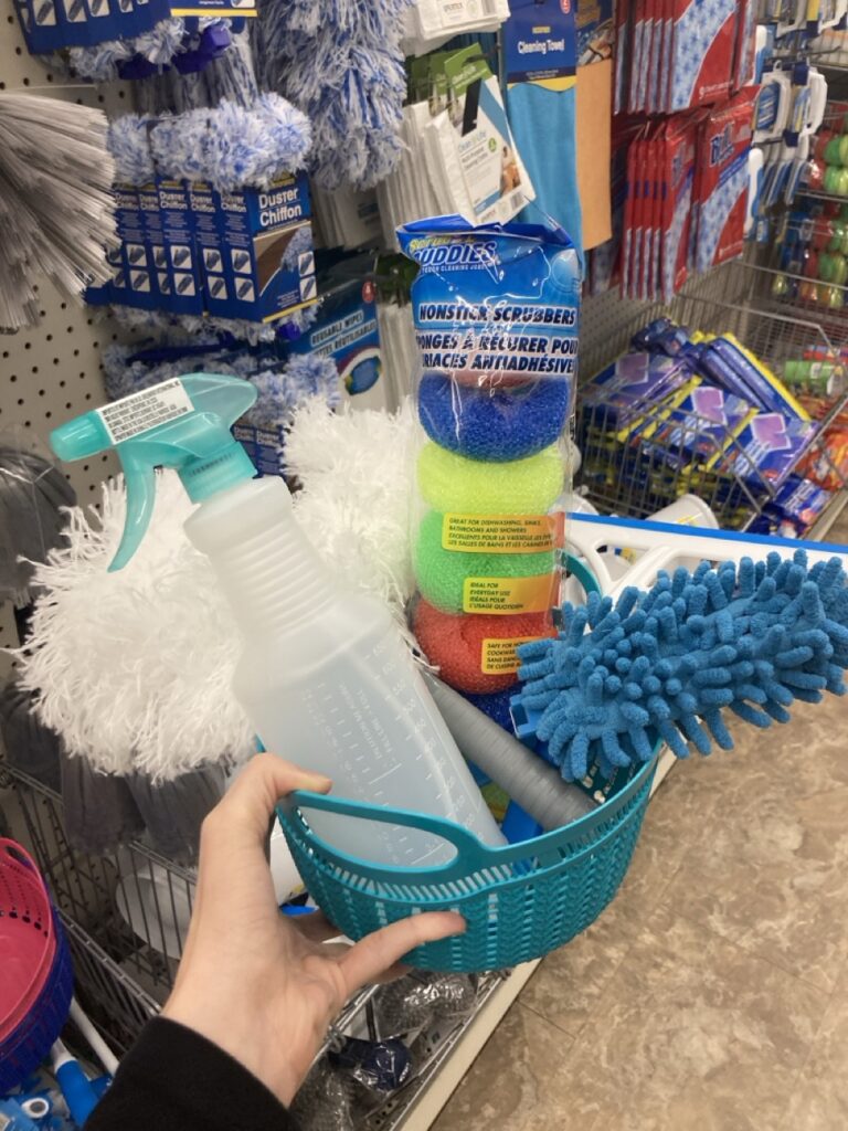 This picture is of a small plastic basket filled with cleaning tools being held by a hand in an aisle at the dollar store