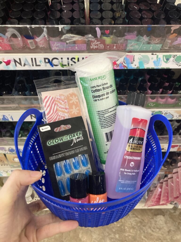 This picture is of a small plastic basket filled with nail polish and nail accessories being held by a hand in an aisle at the dollar store