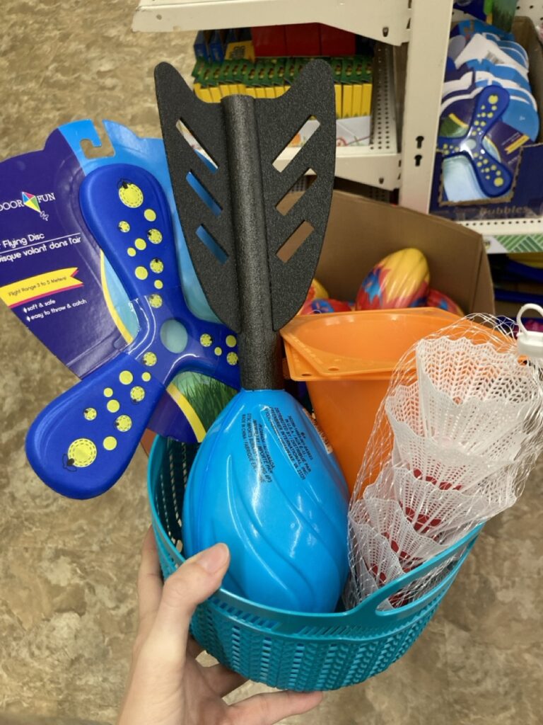 This picture is of a small plastic basket filled with outdoor games and sports toys being held by a hand in an aisle at the dollar store