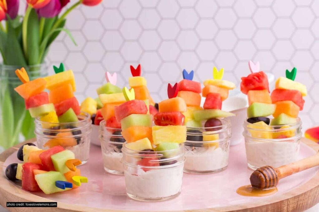 There are little glass jars with kebabs of fruit pieces with a white cottage cheese dip in the base of the jars. 