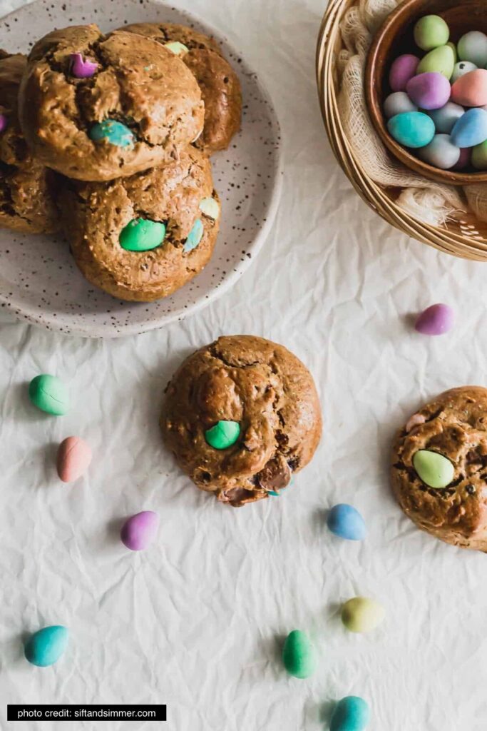 There is a plate of cookies with colorful mini easter eggs in them. There are a few cookies and easter eggs scattered on the table too. 