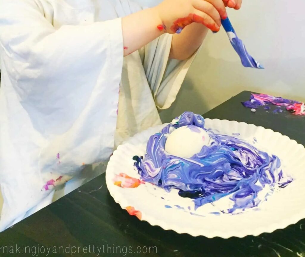 There is a paper plate with shaving cream mixed with food color with a hard boiled egg in it. You can see the hands of a toddler above it with a paint brush. 