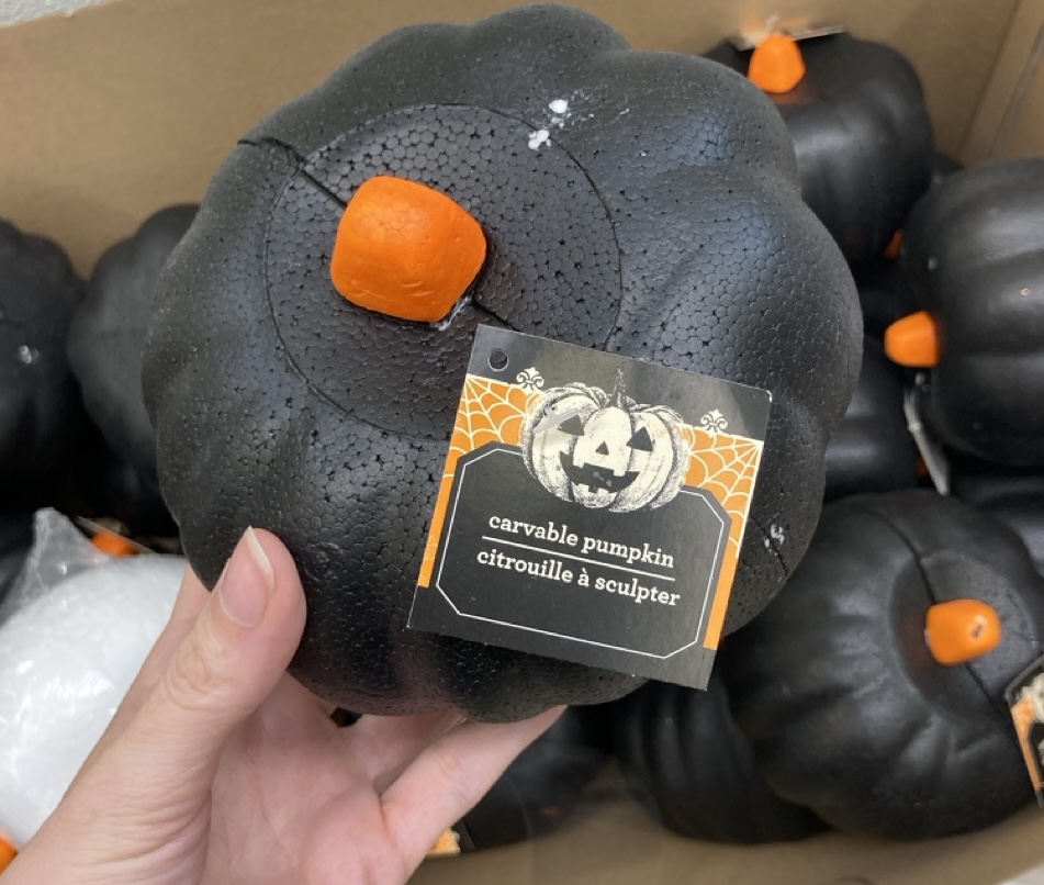 A hand holding a black foam pumpkin with a sticker indicating it is carvable, displayed in a cardboard box with other pumpkins.
