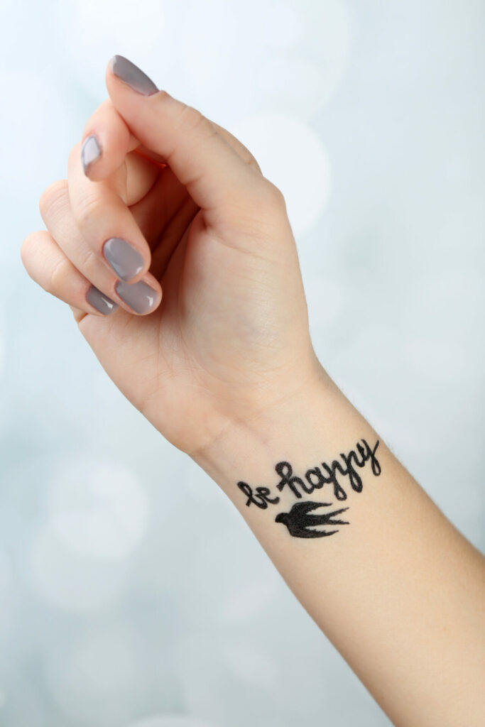 a dark black wrist tattoo that says be happy with a little bird