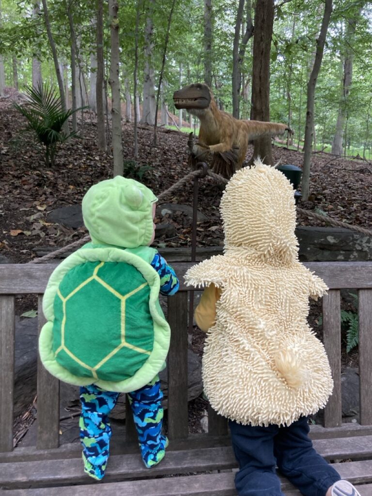 The backs of toddlers in a turtle and duck costume looking onto a dinosaur statute