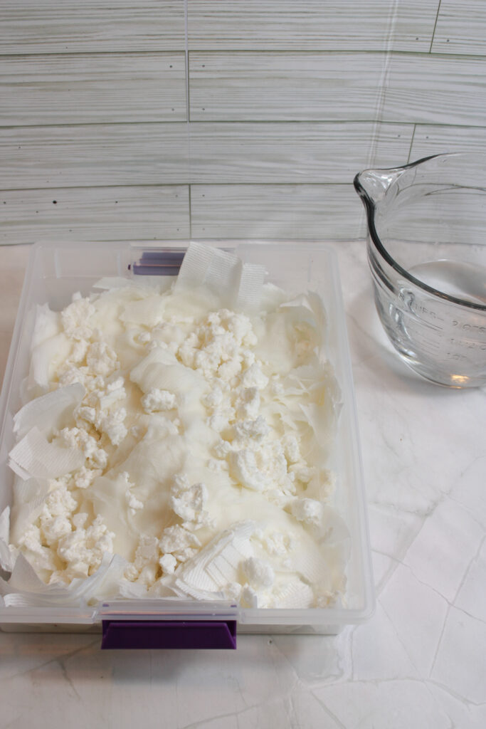 Mixed melted bar of soap and shredded toilet paper inside a clear plastic container and a measuring cup with water on the side.